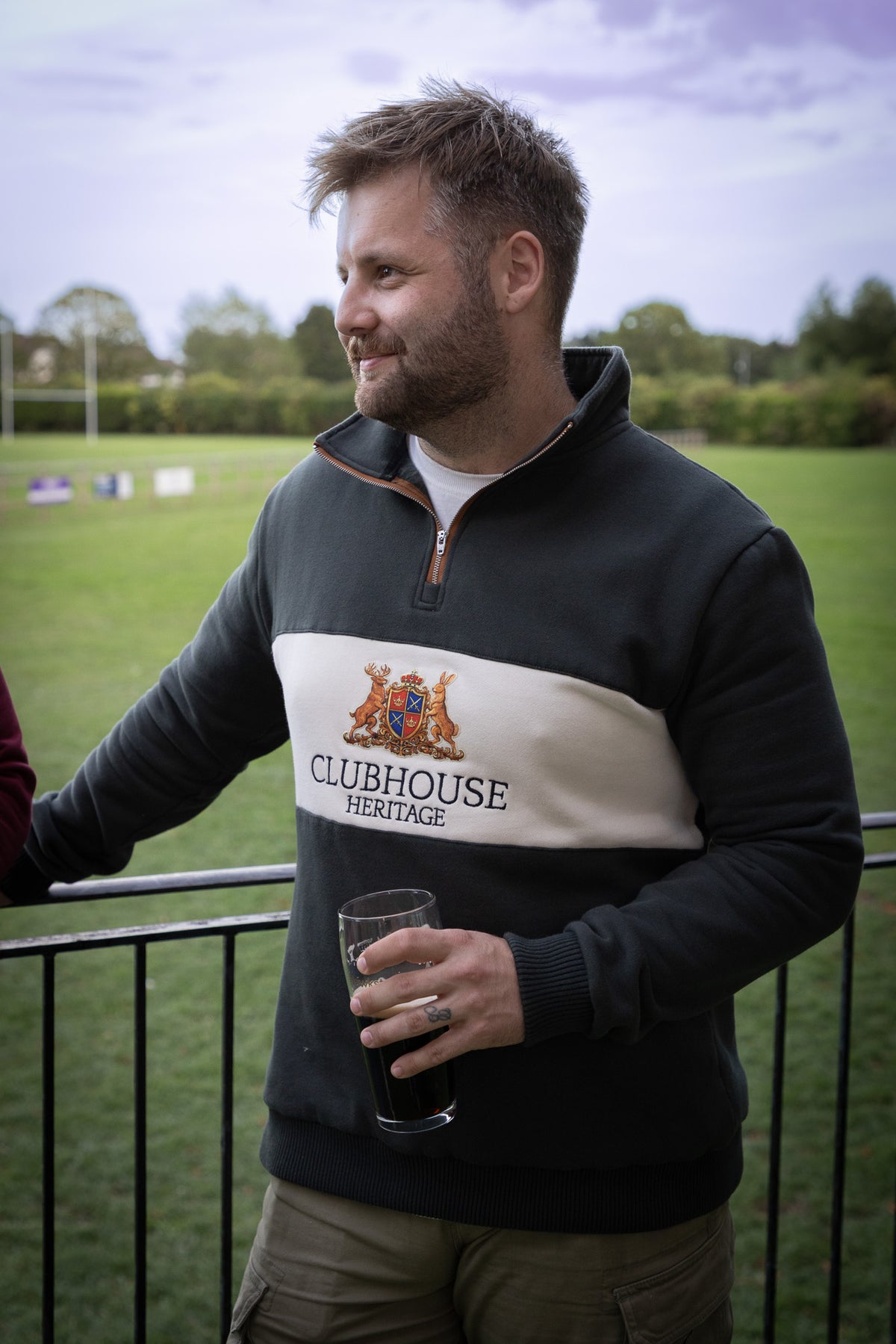 A man wearing a Clubhouse Heritage quarter-zip pullover sweater, standing outdoors on a balcony with a glass of dark beer in hand, with a green field and cloudy sky in the background.