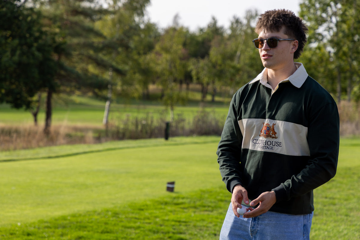 A man wearing a dark green and beige Clubhouse Performance long-sleeve rugby shirt stands on a grassy golf course wearing sunglasses.