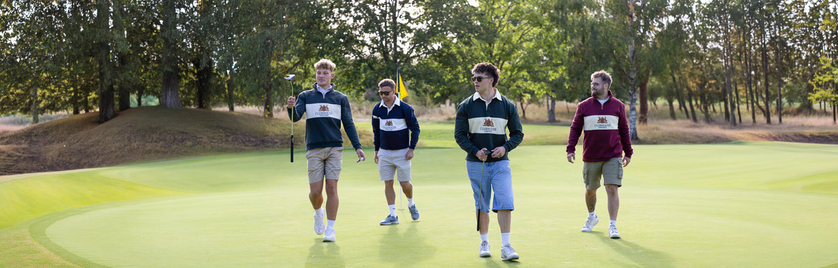 Four men walking together on a golf green, wearing matching dark athletic shirts with a white logo panel on the chest and khaki or maroon shorts.