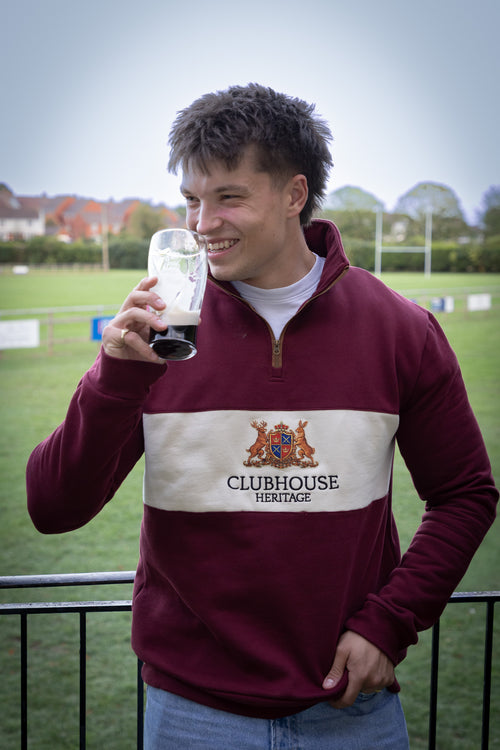 A man wearing a maroon and cream Clubhouse Heritage quarter-zip sweatshirt, holding a pint of stout in a glass, standing on a balcony with a golf course in the background.