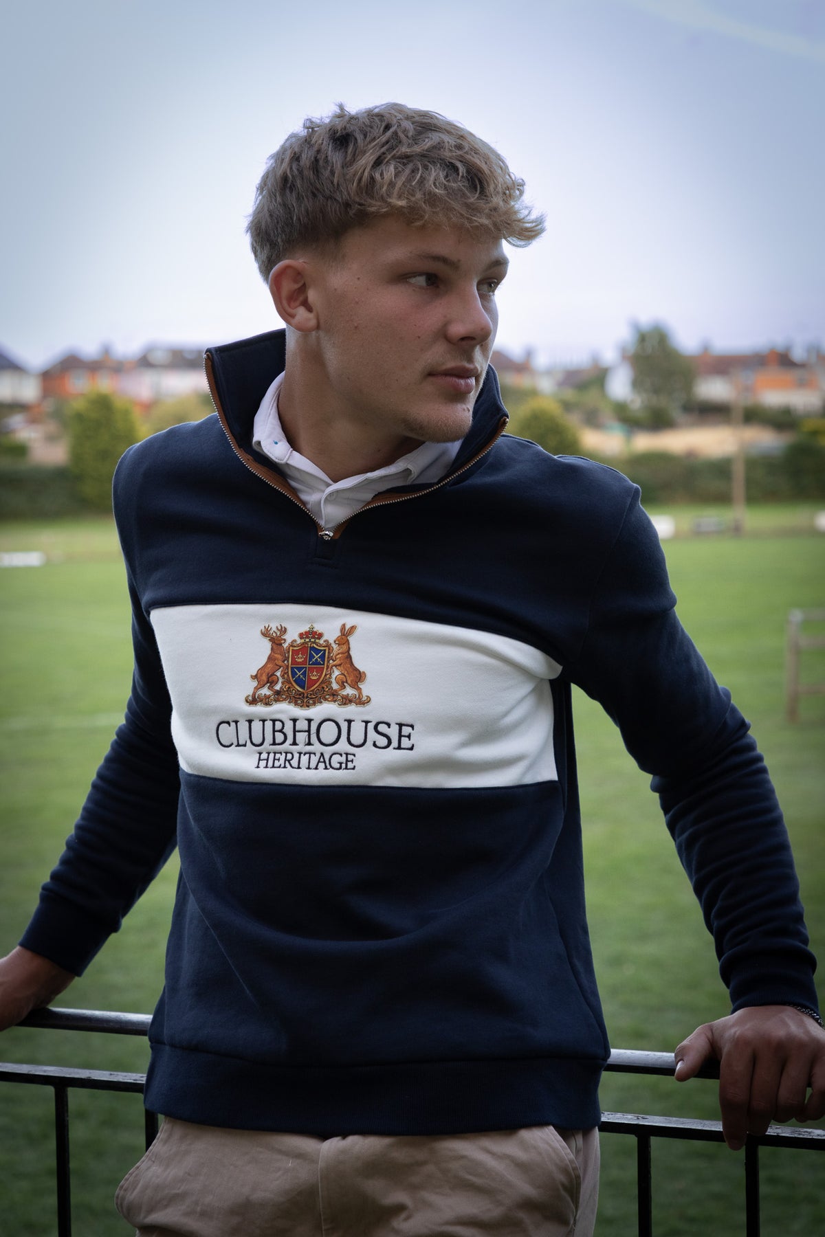 A young man wearing a navy blue and white Clubhouse Heritage quarter-zip sweatshirt, standing outdoors on a grassy field with a fence in the foreground.