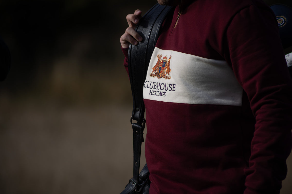A close-up of a person wearing a maroon long-sleeve shirt with a wide white horizontal stripe across the chest. The stripe features a crest and the text 'CLUBHOUSE HERITAGE'.